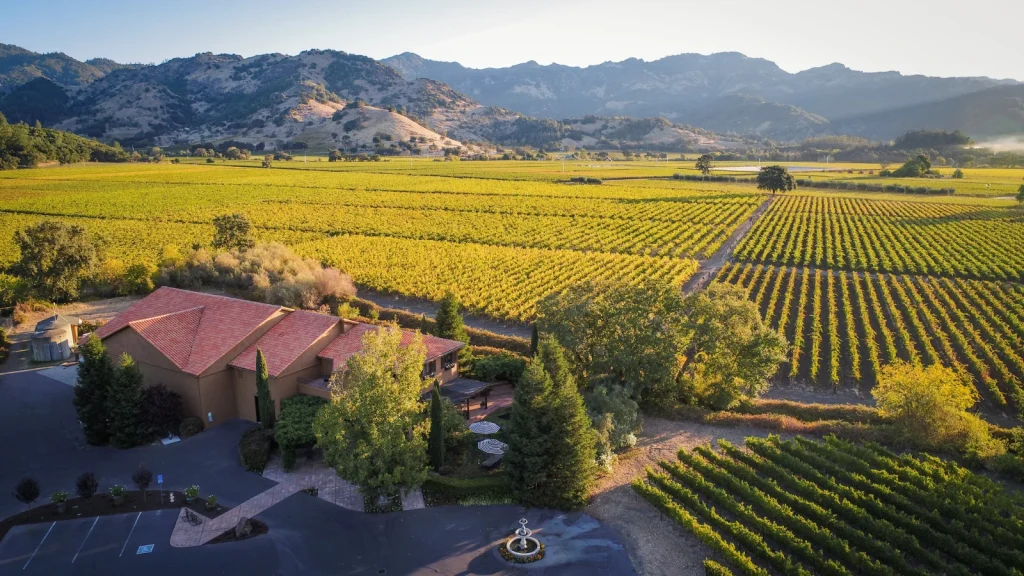 Aerial view of Bennett Lane Winery showcasing the estate buildings surrounded by expansive vineyard rows stretching across the valley floor with dramatic mountain ranges in the background during golden hour