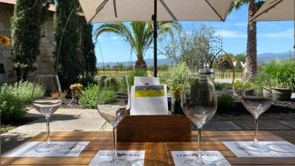 Elegant wine tasting setup at Hagafen Cellars featuring multiple wine glasses arranged on tasting sheets with vineyard views and umbrellas providing shade on the outdoor patio