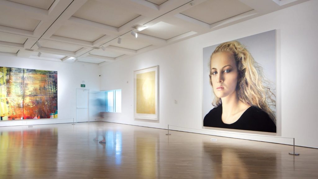 Interior gallery space at Hess Persson Estates Museum featuring contemporary art exhibitions including abstract paintings and a large portrait displayed on white walls with polished concrete floors.