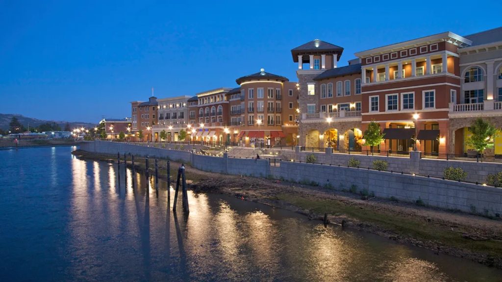 Napa Riverwalk's elegant mixed-use buildings illuminated at twilight with their warm lights reflecting in the Napa River waters along the scenic waterfront promenade