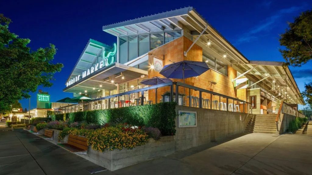Oxbow Public Market's modern glass building illuminated at dusk with outdoor dining umbrellas and landscaped planters creating an inviting evening atmosphere in Napa Valley