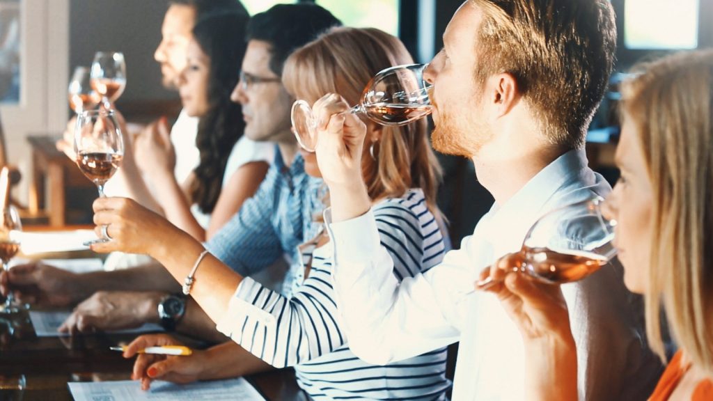 Group of friends enjoying a social wine tasting in NAPA with a man in the foreground savoring red wine while others participate in the tasting experience at a vineyard