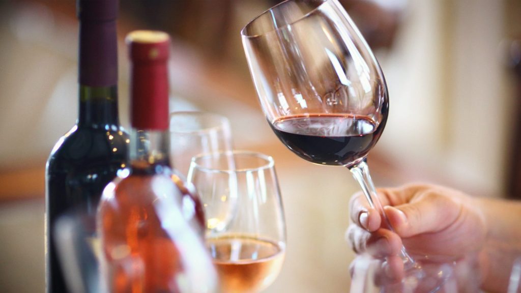 Professional wine tasting in NAPA featuring a hand holding a glass of red wine with wine bottles and additional tasting glasses in the background at a vineyard setting