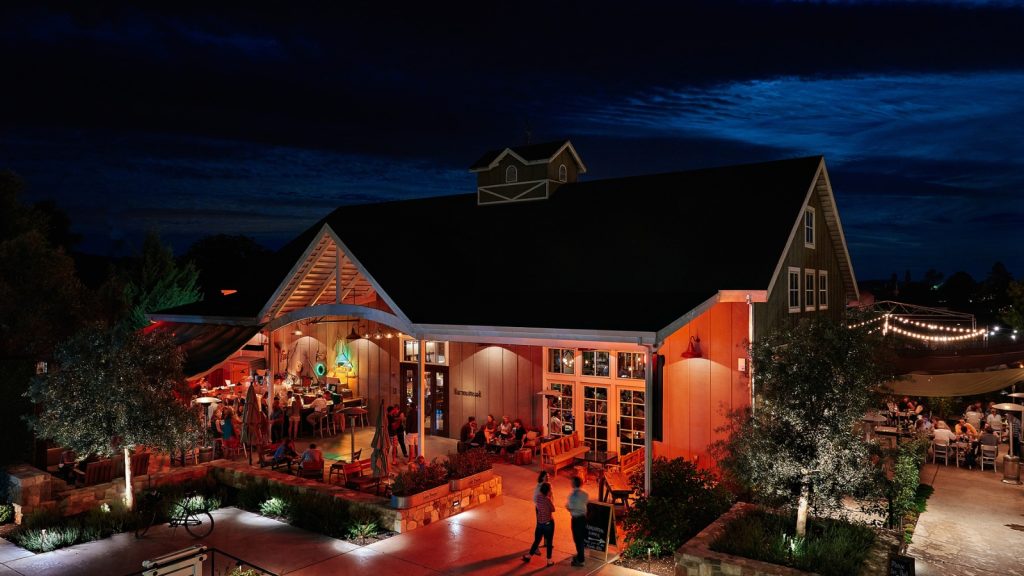 Evening view of Long Meadow Ranch hospitality complex featuring warmly lit restaurant buildings with outdoor dining patios, string lighting, guests dining al fresco, and landscaped grounds creating an inviting farm-to-table dining destination.