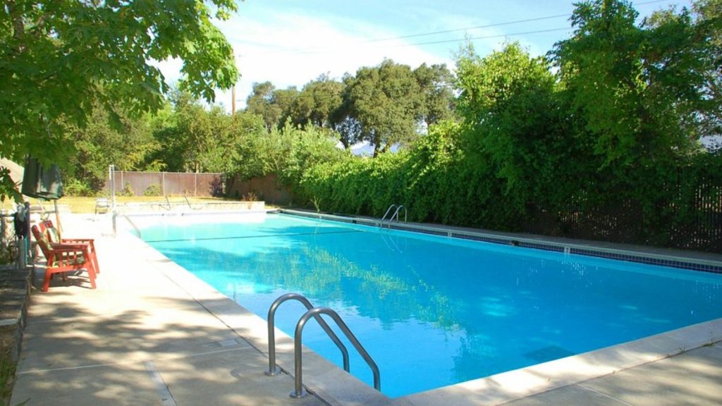 Crystal clear spring-fed pool at Napa Valley State Park featuring bright blue water surrounded by concrete decking, wooden lounge chairs, and mature trees creating a refreshing swimming area in a natural park setting.