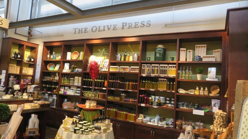 Interior of The Olive Press (Oxbow Public Market) featuring extensive wooden shelving displays filled with olive oils, vinegars, and gourmet products, with a central tasting table and specialty food items in a well-lit retail space.