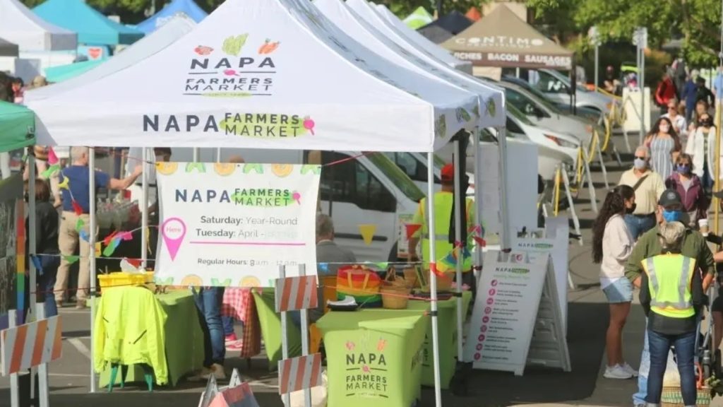 Crowded Napa Farmers Market information booth showing market hours and schedule with shoppers browsing vendor stalls in the bustling outdoor marketplace.