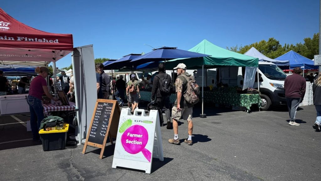 Busy outdoor scene at Napa Farmers Market with vendor tents, shoppers browsing, and a Farmer Section sign visible under blue skies on market day.
