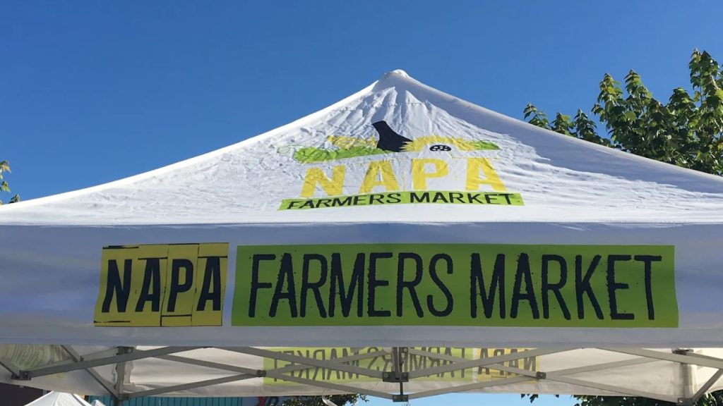 White pop-up tent canopy displaying Napa Farmers Market branding with mountain graphics and clear signage against a bright blue sky at the outdoor market venue.