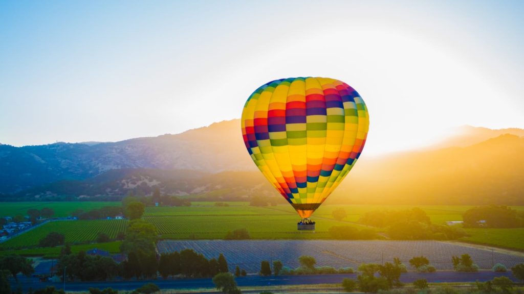 A vibrant rainbow-colored Napa hot air balloon floating above vineyard rows at sunrise with golden light illuminating the valley and surrounding mountains.