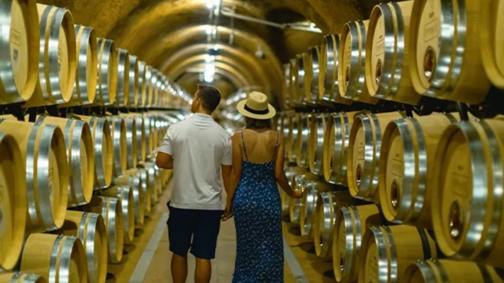 Couple touring Rombauer Vineyards Cave walking through the illuminated barrel room with rows of oak wine barrels extending down the arched underground tunnel.