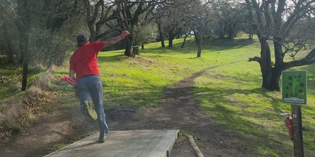 Disc golfer in red shirt throwing from concrete tee pad at Sky Wilderness Park disc golf course, with green fairway winding through bare oak trees and course sign visible on wooded hole.