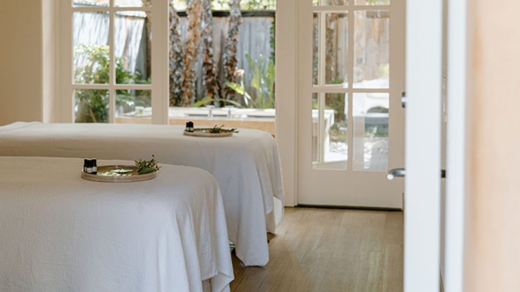 Auberge du Soleil Spa features a serene treatment room with two massage tables draped in white linens, large windows overlooking gardens, and natural daylight ambiance.