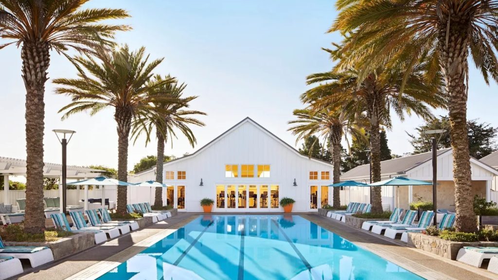 Carneros Resort and Spa showcases a luxurious lap pool lined with lounge chairs flanked by tall palm trees, with a modern white pavilion building and spa facilities visible.