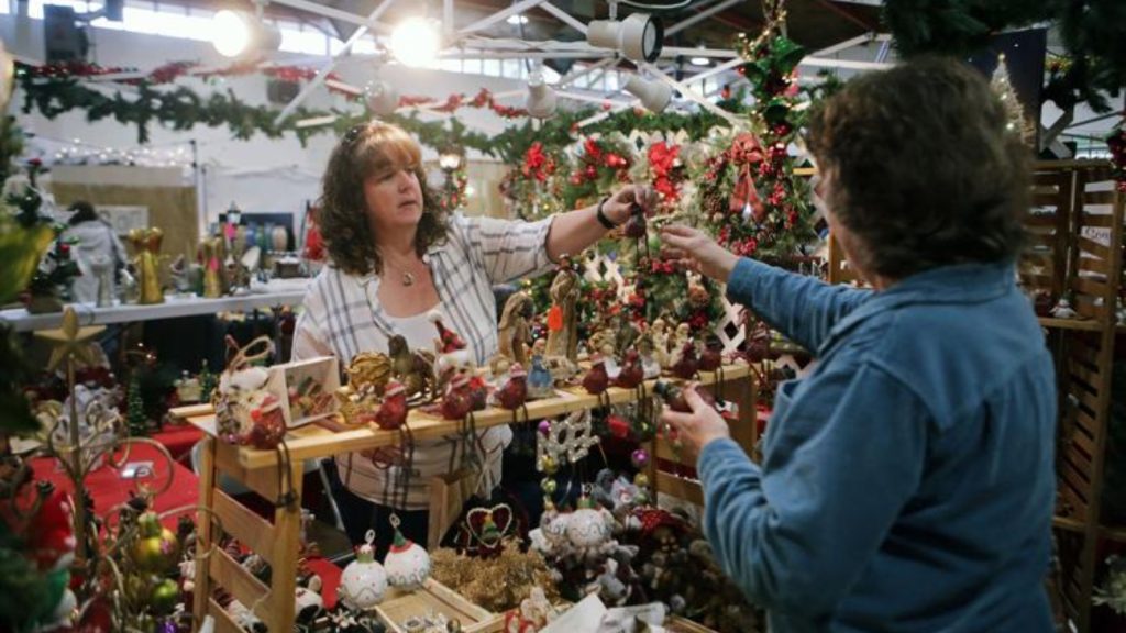 Shoppers browse festive holiday decorations and ornaments at a napa valley christmas market or craft fair, featuring handmade wreaths, garland, and seasonal décor in a warehouse setting.