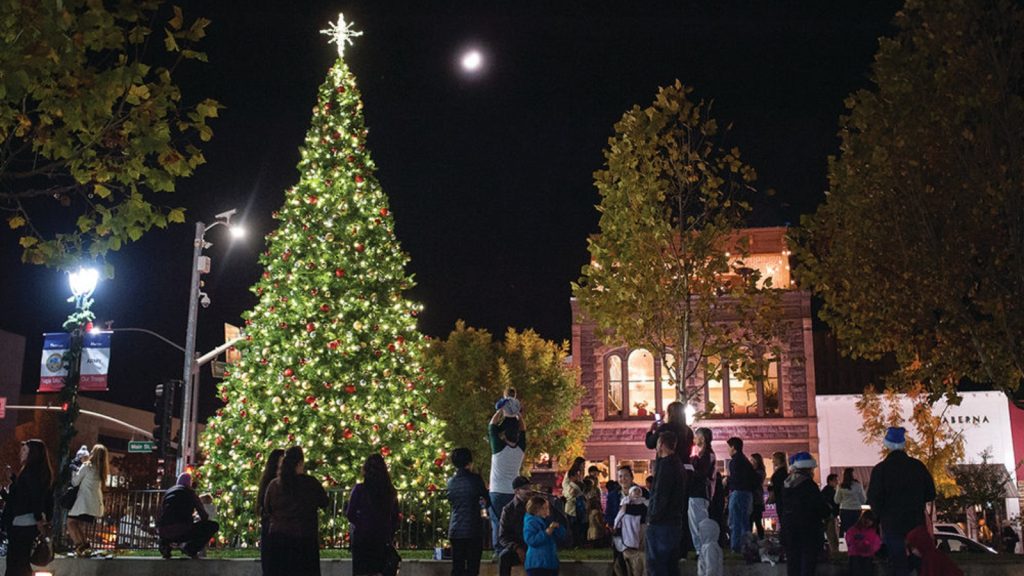 Families gather around a towering illuminated Christmas tree at napa valley christmas celebrations, with historic brick buildings and festive lighting creating a holiday atmosphere under the moonlit night sky.