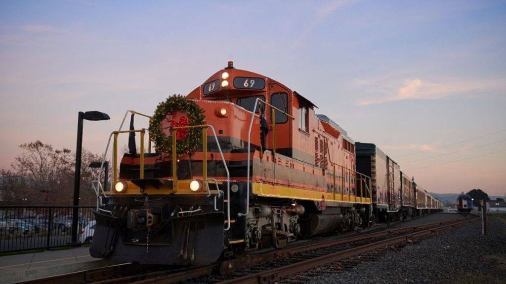 A festive red and yellow locomotive decorated with a holiday wreath represents the Napa Valley Wine Train's Jolly Santa Journey, traveling through scenic wine country during the Christmas season.
