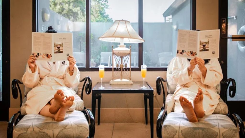 The Spa at Silverado features a relaxation lounge with two guests in spa robes enjoying refreshments, comfortable seating, ambient lighting, and panoramic windows overlooking the grounds.