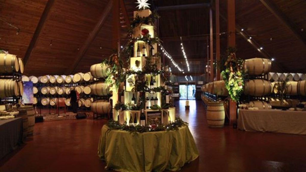 A festively decorated wine barrel structure adorned with holiday garland and lights stands in a barrel room during Winery Christmas Tastings, surrounded by oak barrels and wine tasting stations.