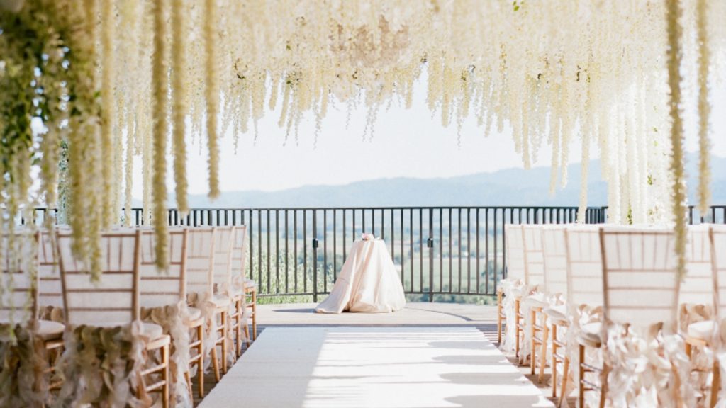 An elegant outdoor wedding ceremony at Auberge du Soleil features cascading wisteria florals, wooden chiavari chairs, and a bride in flowing white gown framed by valley and mountain vistas.