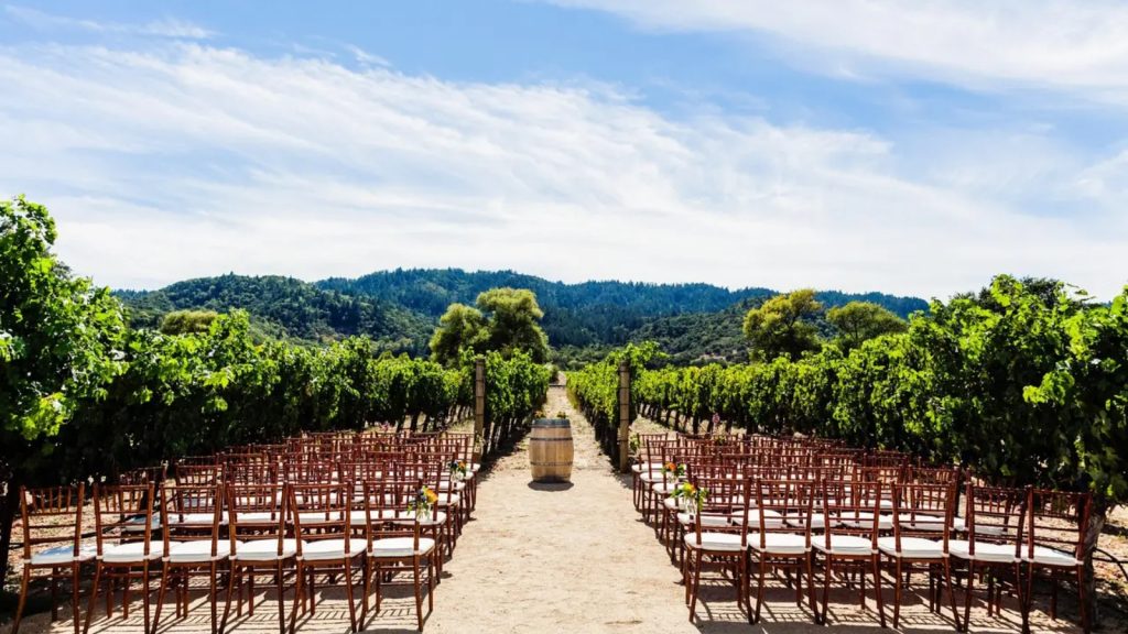 An outdoor wedding ceremony aisle lined with wooden chiavari chairs stretches through lush vineyards at Brix Restaurant & Gardens, with forested hills visible in the distance beneath a blue sky.