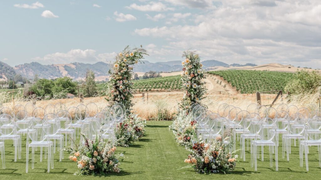 A picturesque outdoor wedding ceremony at Carneros Resort and Spa features white chiavari chairs arranged on manicured lawn, floral-adorned archway, and expansive vineyard views beneath a blue sky with rolling hills.