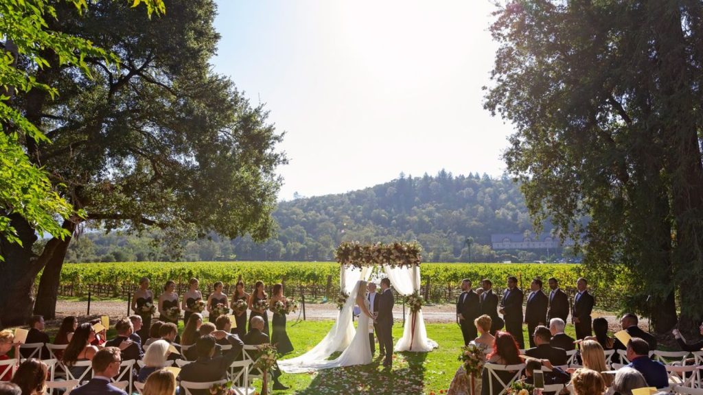 A bride and groom stand beneath a white wedding arch at the altar during an outdoor ceremony at Charles Krug Winery, with guests seated on white chairs framed by ancient oak trees and expansive vineyard views.