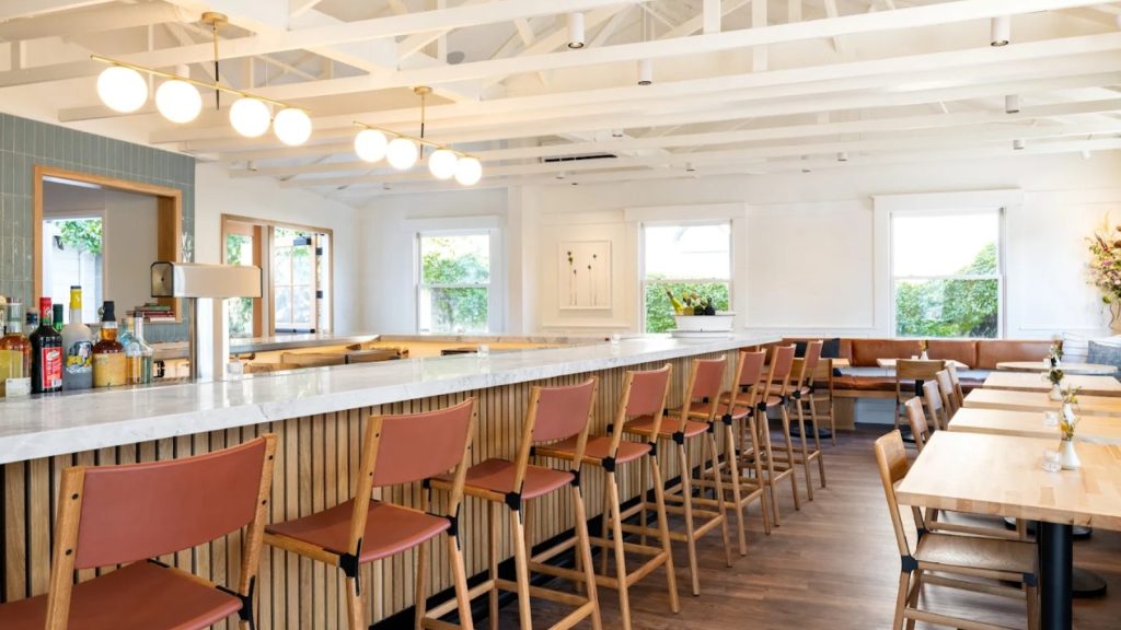Charlie's Napa Valley features a bright, modern tasting room with a long white marble bar, rust-colored seating, wooden accents, and natural light from large windows overlooking vineyards.