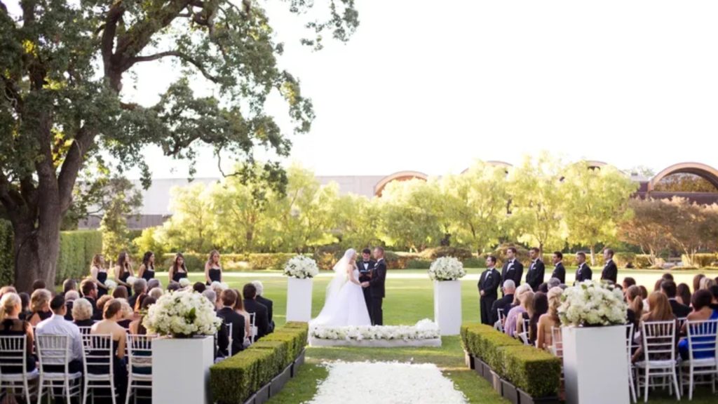 A formal outdoor wedding ceremony at Domaine Chandon features a bride and groom at the altar beneath mature oak trees, white floral arrangements in pedestals, guests seated on chiavari chairs, and manicured gardens with vineyard views.