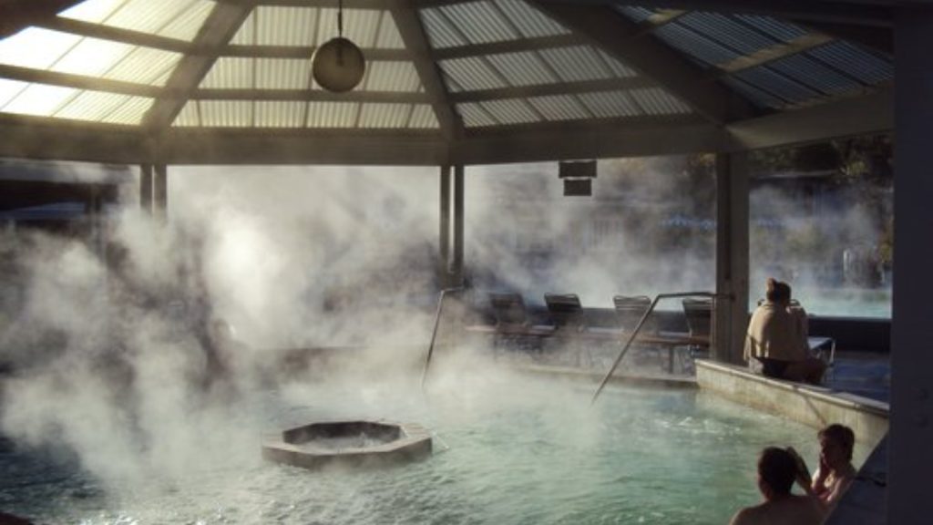 Indian Springs Calistoga guests relax in a steaming geothermal hot spring pool surrounded by mineral-rich vapor under a modern covered pavilion structure.