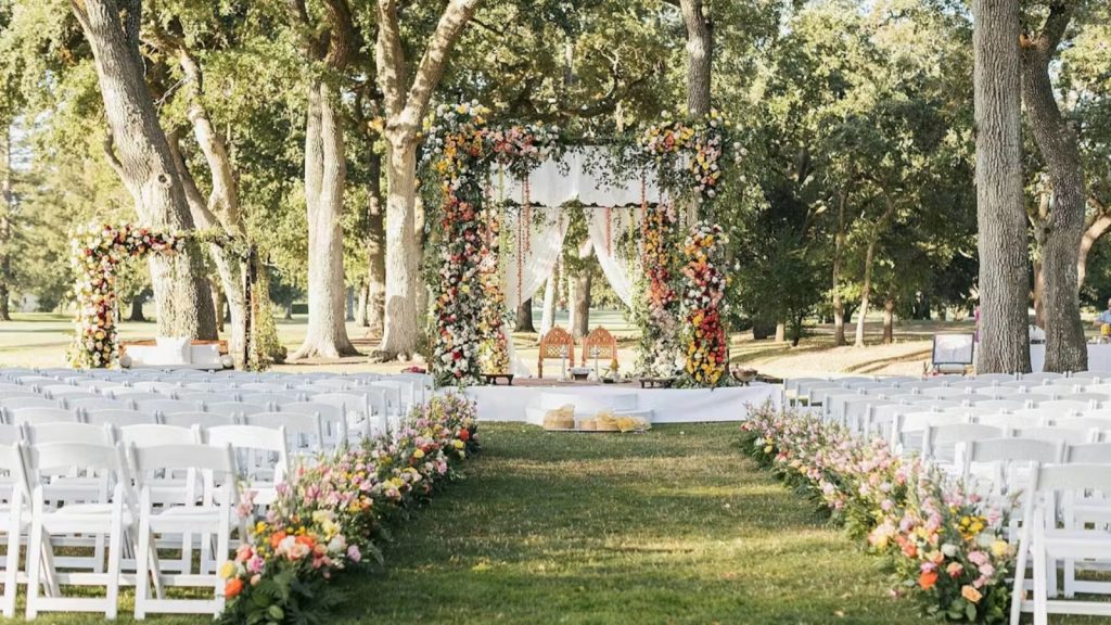 An elegant outdoor wedding ceremony setup at Silverado Resort features a floral-adorned arch beneath towering oak trees, white folding chairs lining the aisle, and colorful flower arrangements in an expansive manicured lawn setting.