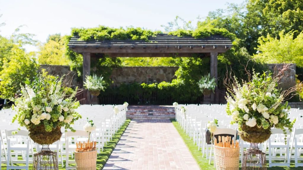 An elegant outdoor wedding ceremony at The Estate Yountville features white chiavari chairs lining a brick aisle, abundant white and green floral arrangements in urns, a modern pergola structure, and lush landscaping beneath overhanging trees.