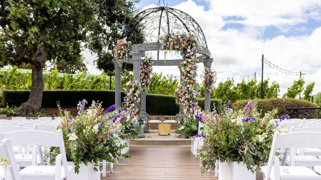 A stunning wedding ceremony setup at The Meritage Resort & Spa features a decorative gazebo with floral arrangements, white folding chairs lining a wooden aisle, and vibrant purple and pink flowers overlooking vineyard views.