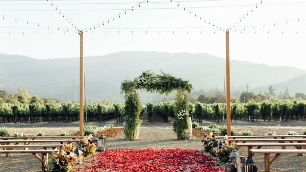 An outdoor wedding ceremony setup at Tre Posti features a green-draped arch decorated with white flowers, wooden bench seating, red petal aisle, string lights overhead, and sweeping vineyard and mountain views.