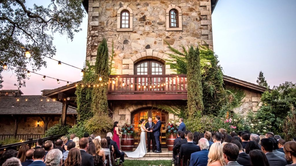 A bride and groom exchange vows at the altar in front of a historic stone castle tower at V. Sattui Winery, with wedding guests seated on the lawn during an elegant twilight ceremony.