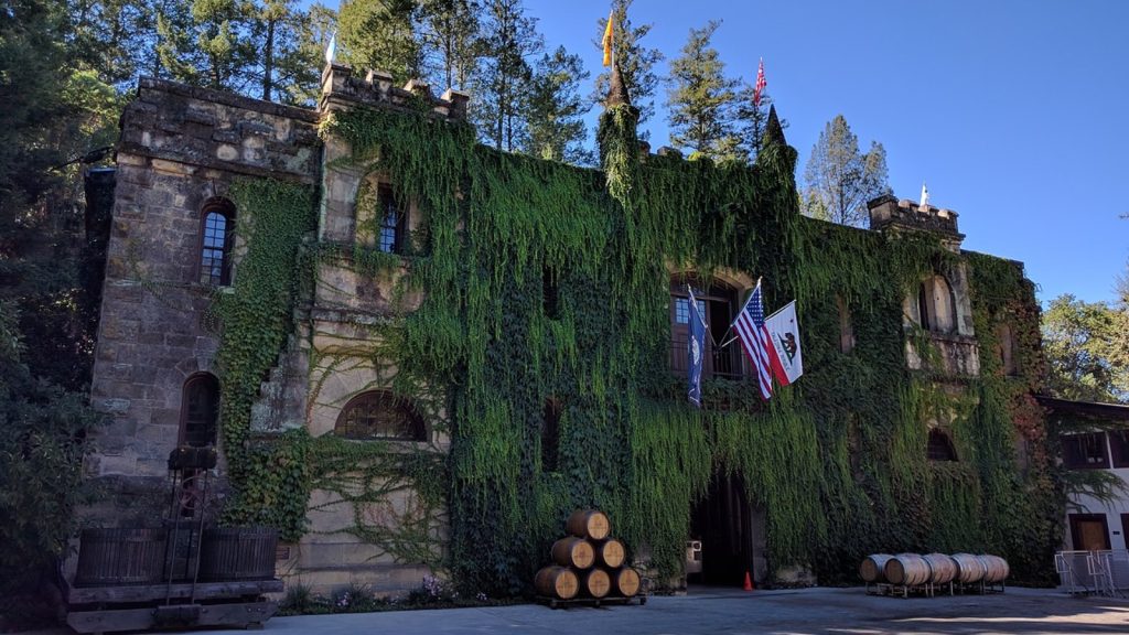 Chateau Montelena stone castle facade covered in ivy with American flag, wine barrels, and trees under clear blue sky