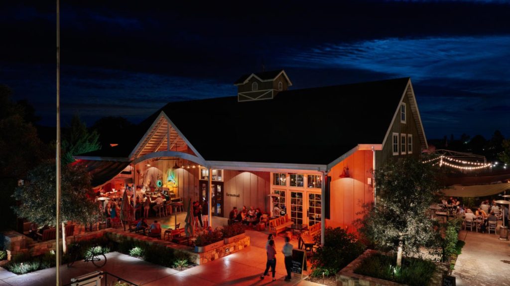Farmstead restaurant illuminated at twilight with warm orange lighting, outdoor dining patios, guests, and string lights against dark blue sky