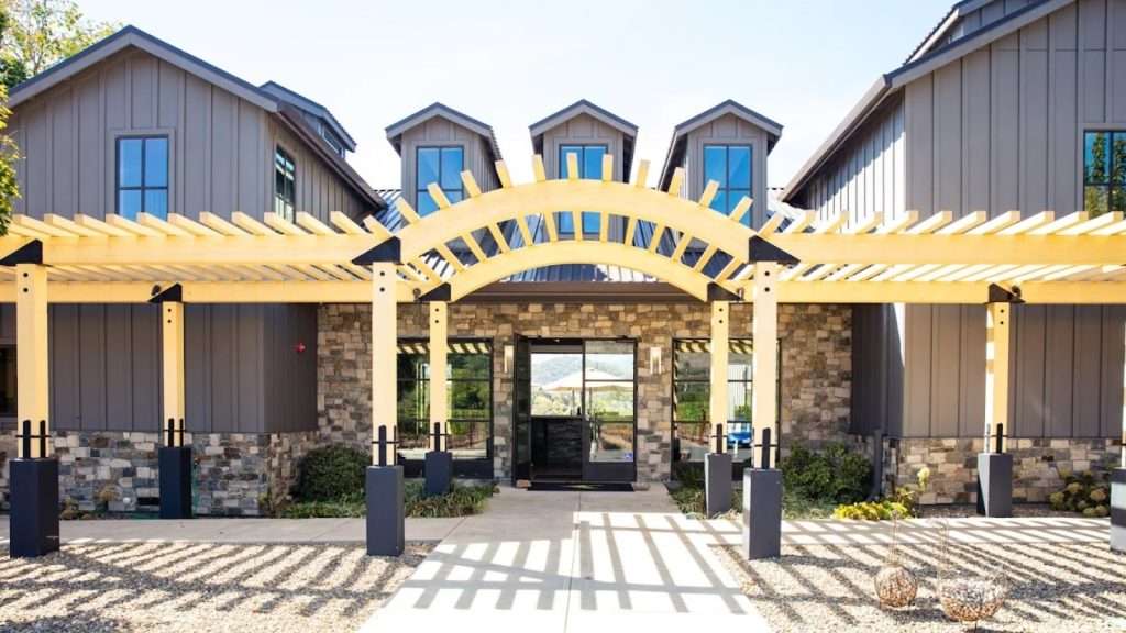 Goosecross Cellars features a contemporary stone and wood tasting facility with a striking yellow pergola archway, modern architecture, and manicured landscaping creating an inviting wine country entrance.
