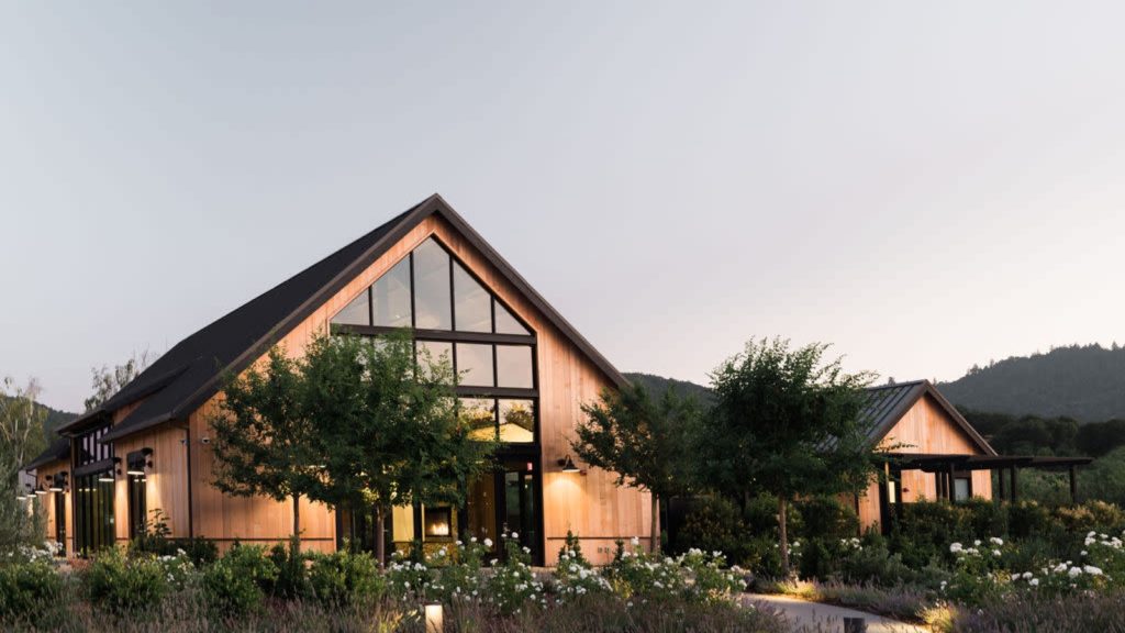 The Estate Yountville showcases modern architectural buildings with natural wood finishes, large windows, surrounding vineyard landscape, and evening lighting in the Napa Valley hills.
