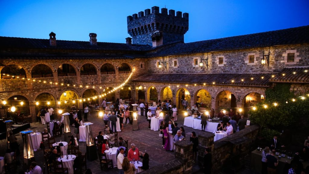 Castello di Amorosa evening event in castle courtyard with stone arches, tower, string lights, cocktail tables, and guests at twilight