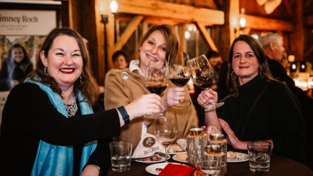 Celebration of Women in Wine Events featuring three smiling women toasting wine glasses at rustic winery dinner with warm lighting and wooden beams