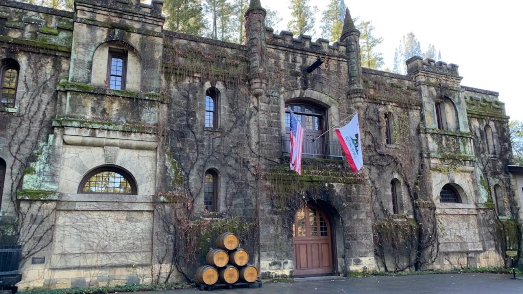 Chateau Montelena weathered stone castle facade with ivy-covered walls, American and California flags, arched doorway, and stacked wine barrels