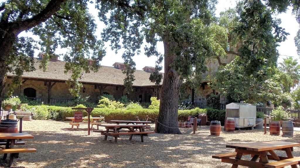 V. Sattui Winery outdoor picnic area with wooden tables, large shade tree, wine barrels, and stone building surrounded by greenery