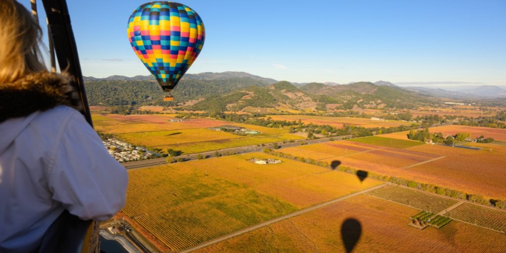 Hot air balloon ride cost - an aerial perspective from inside a hot air balloon basket showing a checkered balloon envelope above orange agricultural fields and green forested mountains with distant valley landscape during daytime flight experience