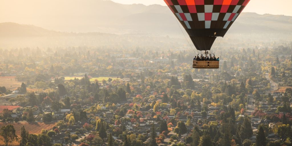 Hot air balloon weather - a red and white checkered hot air balloon with wicker basket suspended above a sprawling urban landscape with mountains in the distance during golden hour sunrise conditions with misty atmospheric weather
