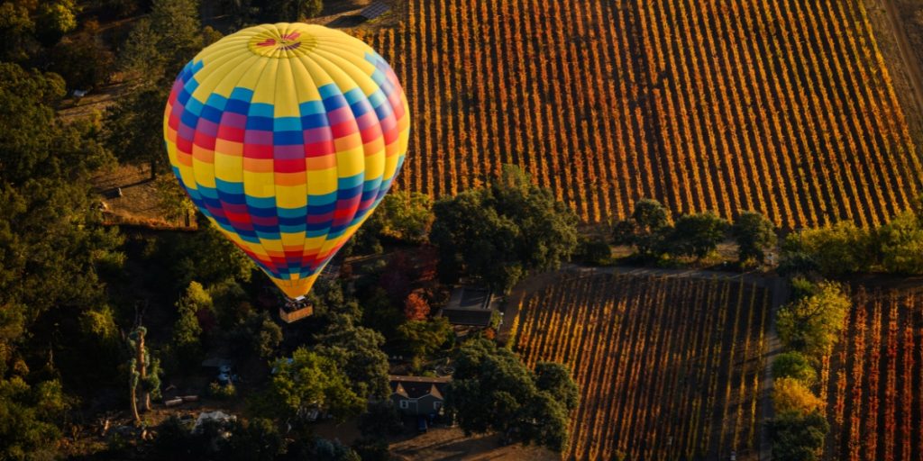 Hot air balloon weather - a colorful yellow, red, and blue striped hot air balloon drifting low over golden vineyard fields and green trees during ideal morning flight conditions with perfect weather visibility