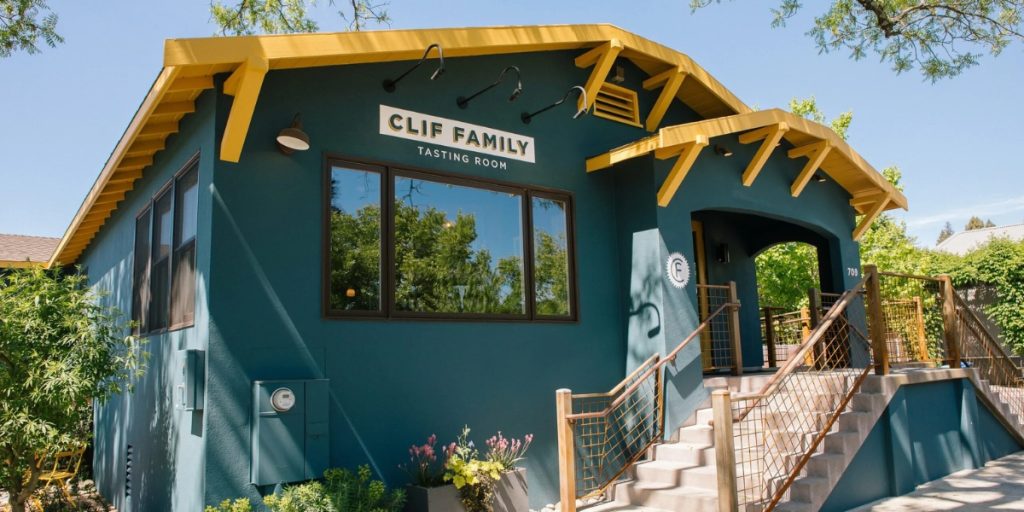 A modern teal and gold Clif Family Tasting Room building featuring contemporary architecture with large windows, wooden beams, and an exterior staircase surrounded by vineyard landscaping in Napa Valley.