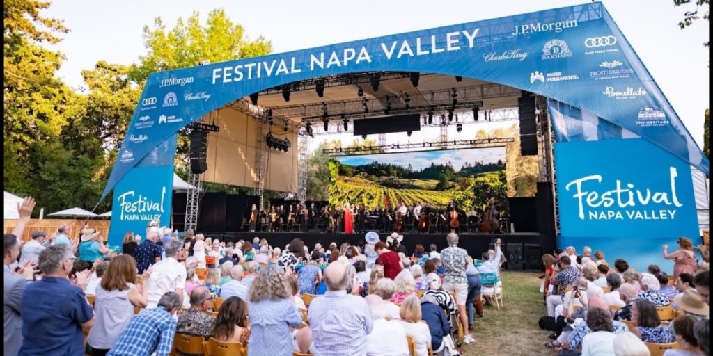 A large outdoor stage at Festival Napa Valley 2026 during Napa July with a crowd of attendees seated on the lawn, featuring sponsor logos and a scenic vineyard backdrop for live performances.
