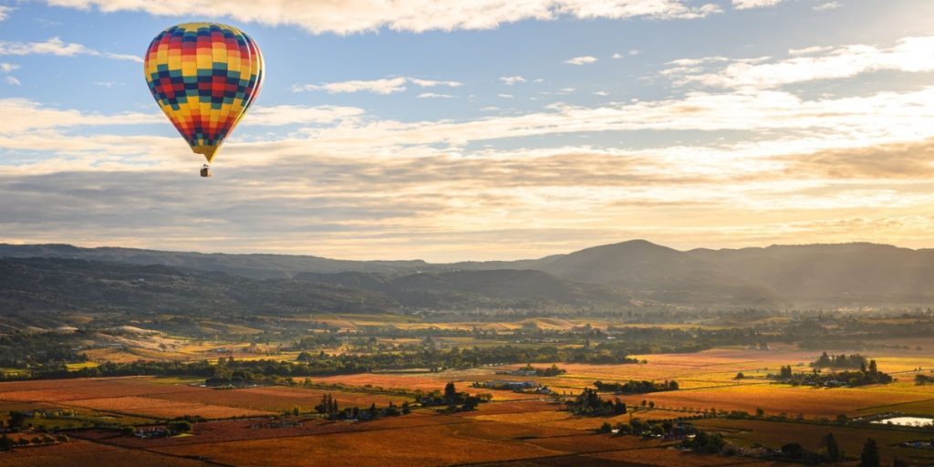 A colorful hot air balloon floating above the scenic Napa Valley landscape during Napa July, showcasing the picturesque rolling hills, vineyards, and golden fields characteristic of Napa Valley Aloft experiences.
