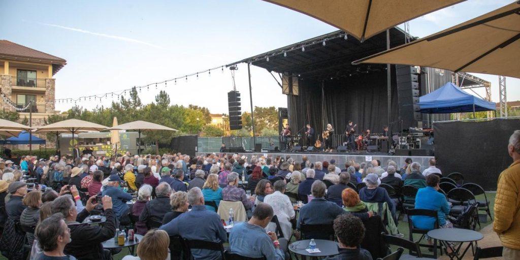 A large outdoor audience enjoying Napa July's summer concerts and live music performances on a shaded stage area with tan umbrellas, seating arrangements, and scenic vineyard views in the background.